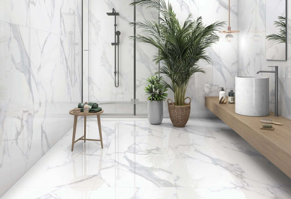 
A modern bathroom features sleek white marble flooring and walls accentuated by elegant faucets. A glass shower enclosure streams water effortlessly in the corner. A wooden vanity holds a round sink, with a large plant nearby. Tiles paired with green towels on a small stool add a natural touch.